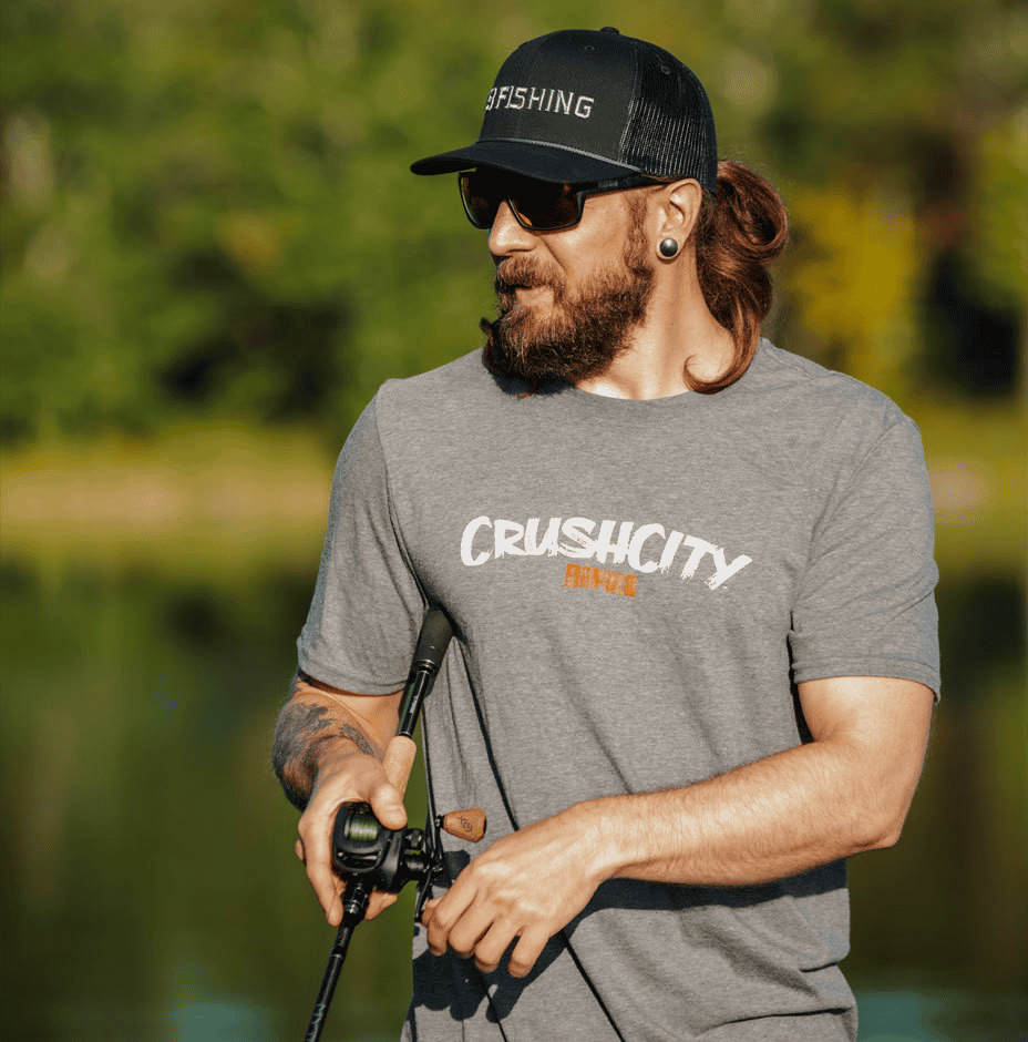 CrushCity Logo T-Shirt 4 A bearded man in sunglasses stands by a lakeside holding a fishing rod, wearing the CrushCity Logo T-Shirt - XL VMC 4 in gray and a black “13 Fishing” cap, with blurred trees and water in the background.