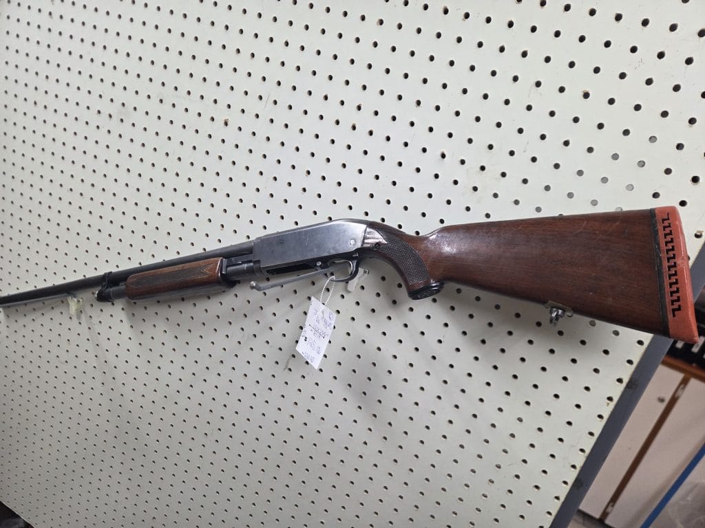 1000014609 pump-action shotgun with a wooden stock and forend mounted on a pegboard wall.
