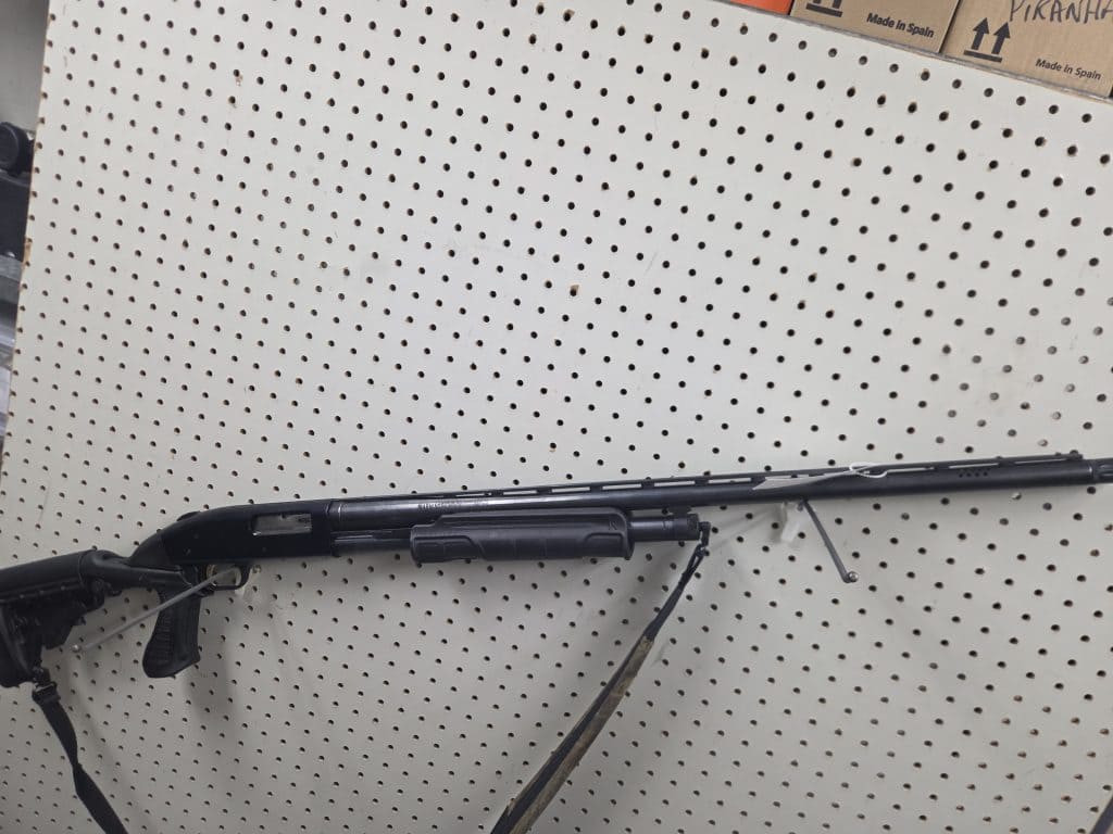 Black 20261018_091035 pump-action shotgun with a tactical stock and shoulder sling mounted on a white pegboard wall.