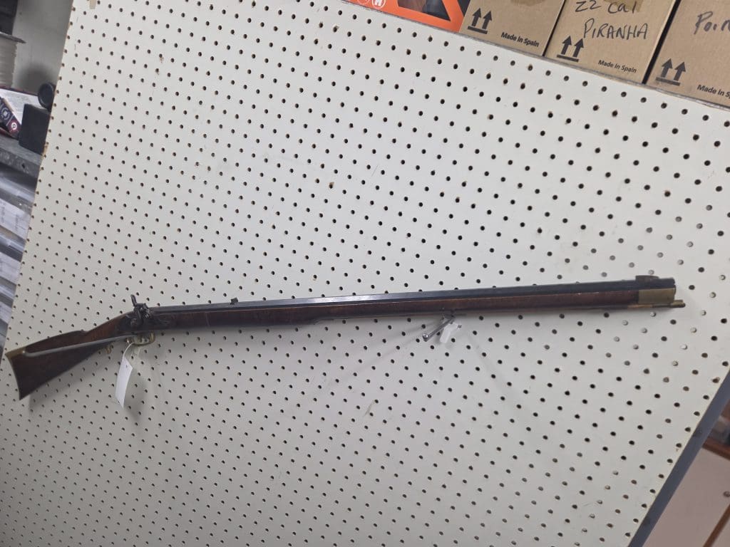 A 20261018_092751 long rifle with a wooden stock and dark metal barrel mounted on a white pegboard display wall.