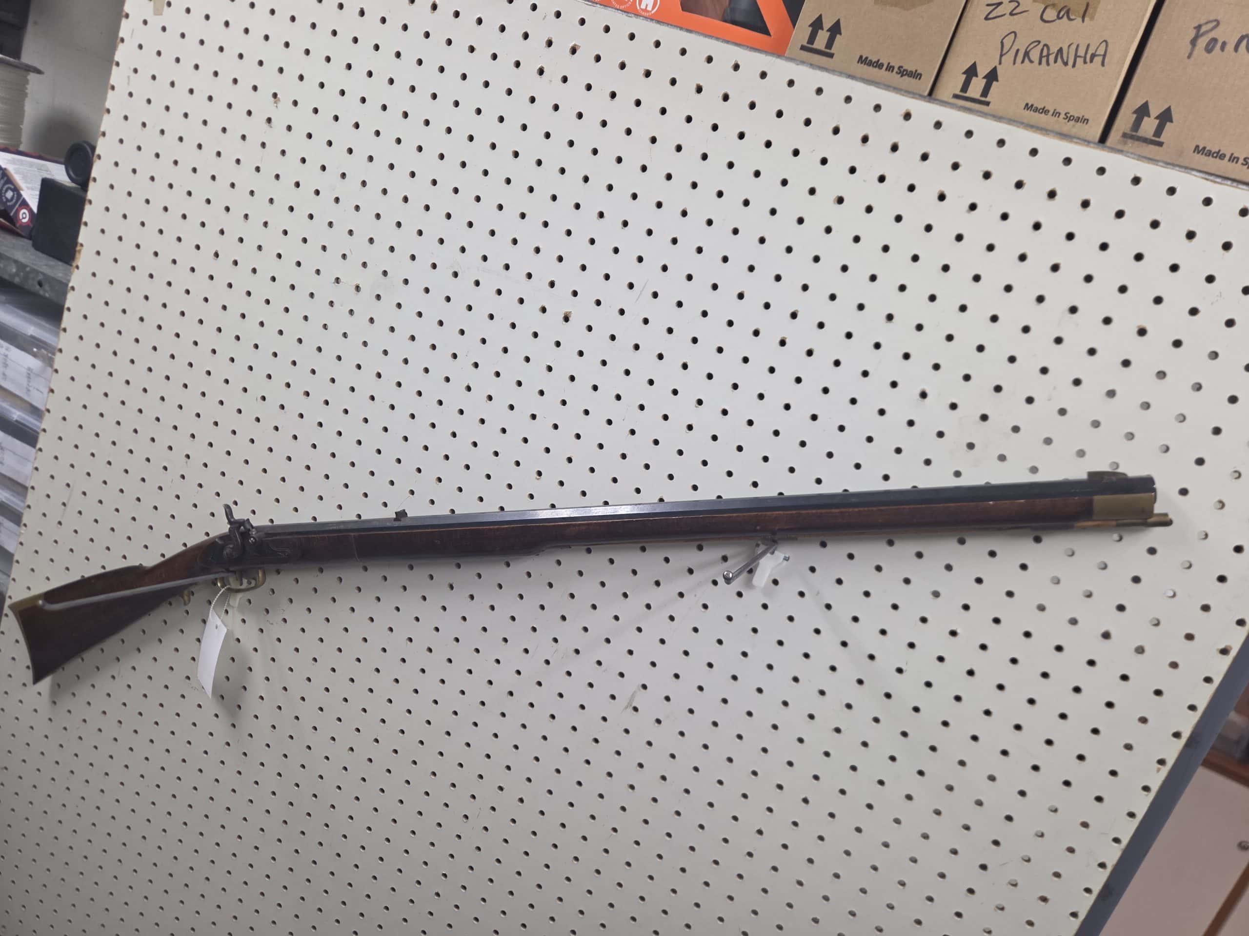 Ardesa 1 A 20261018_092751 long rifle with a wooden stock and dark metal barrel mounted on a white pegboard display wall.