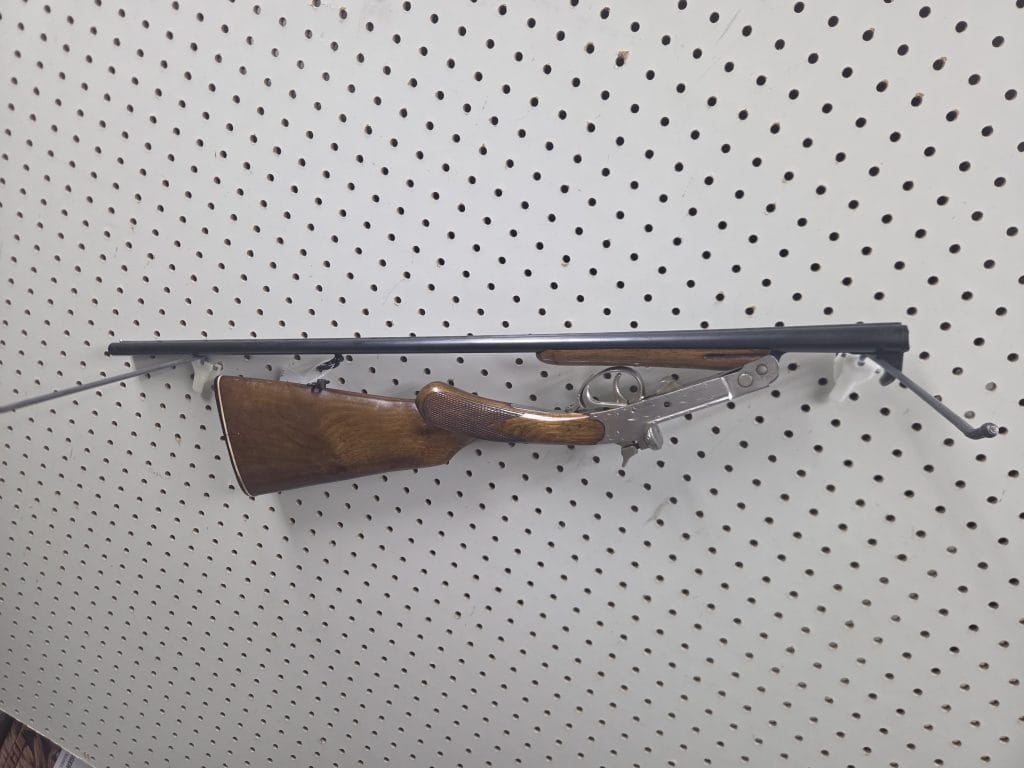 Break-action shotgun 20261018_093014 with a wooden stock mounted on a pegboard wall.