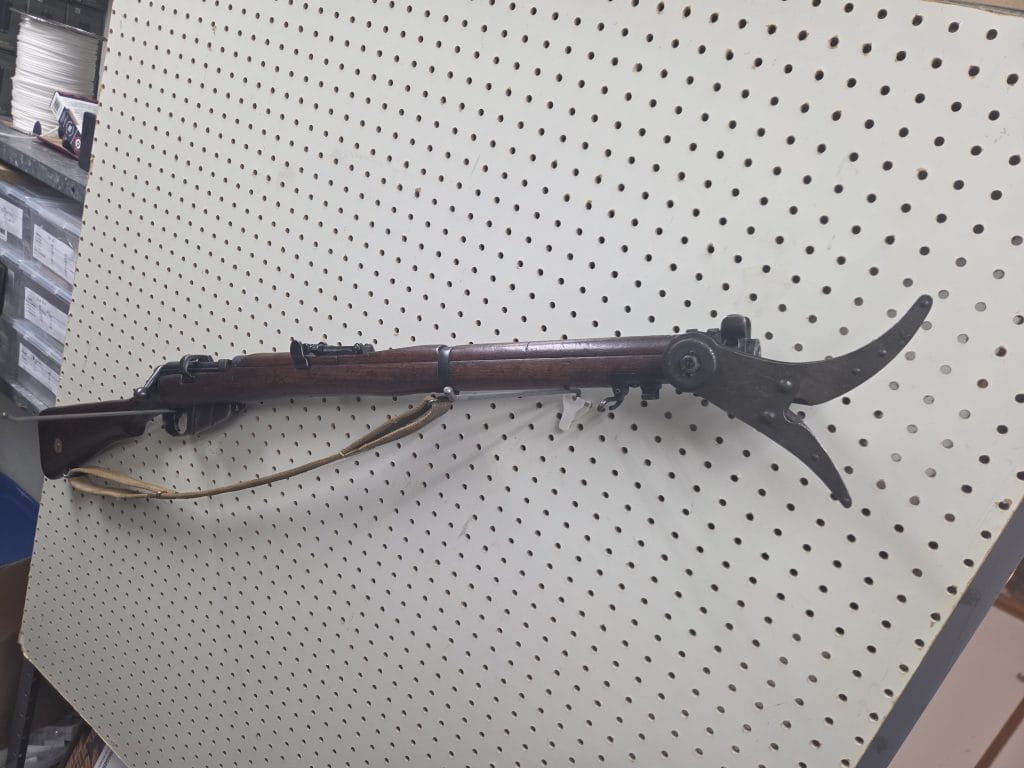 20261018_115424 bolt-action rifle with a tan sling mounted on a pegboard wall, featuring a specialized pronged attachment at the muzzle.